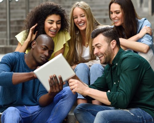 Multi-ethnic group of young people looking at a tablet computer outdoors in urban background. Group of men and woman sitting together on steps.